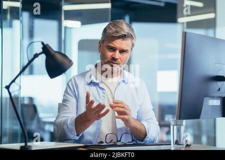 Successful businessman works at the computer, sits at the table and looks at the camera, communicates, explains and gestures Stock Photo