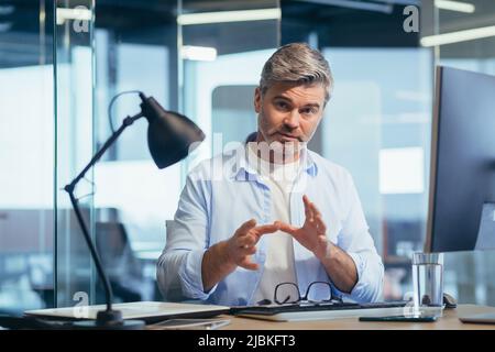 Successful businessman works at the computer, sits at the table and looks at the camera, communicates, explains and gestures Stock Photo