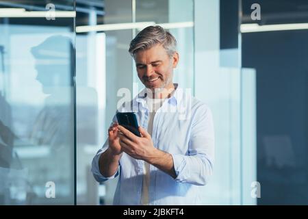 Senior boss business man in modern office, portrait of business owner using phone smiling and rejoicing Stock Photo
