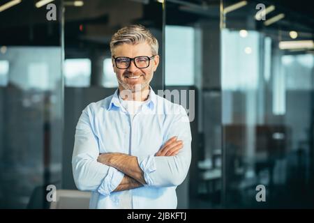 Portrait of senior and experienced male boss boss, with arms crossed, smiling and looking at camera, businessman with glasses, and modern office Stock Photo