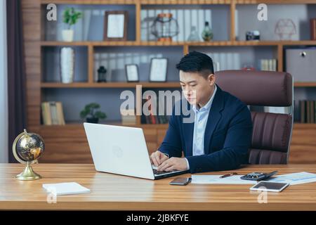 Young handsome male asian businessman, lawyer, working on laptop in the office, sitting at the table in a black suit, pensive Stock Photo