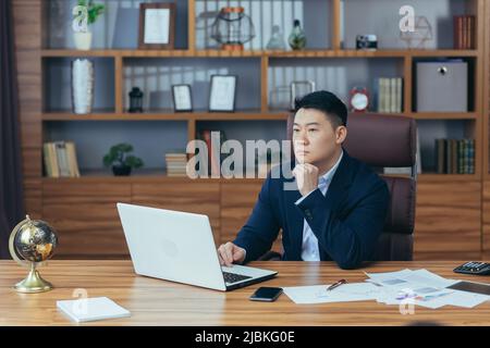 Pensive, thoughtful, worried young Asian man, banker, businessman, director. He is sitting at his desk in his office, working on a laptop Stock Photo