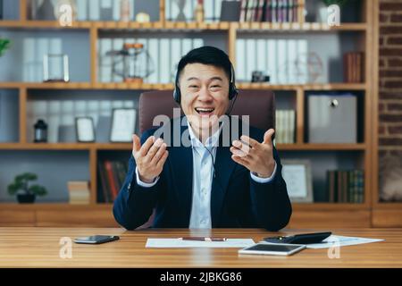 Happy Asian businessman boss looks at the camera smiling and talking to a colleague in an online conference, uses a headset, a man in a business suit Stock Photo