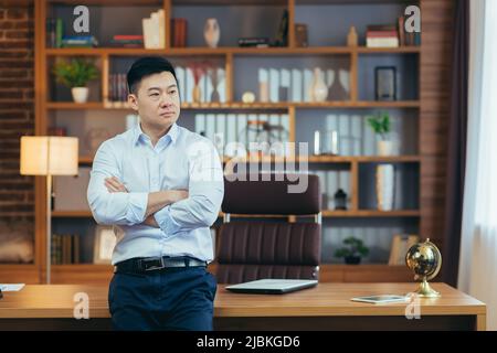 Portrait of a pensive businessman, a man with arms crossed in a classroom office working, looking out the reverse window, Asian thinks Stock Photo