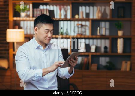 Businessman working on tablet in the evening, man looks at the screen and smiles, works late in the office, happy asian corresponded on a dating site Stock Photo