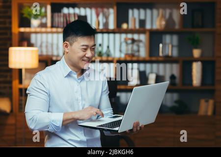 Asian at home in the evening, standing and watching videos online on laptop, man smiling, resting after work Stock Photo