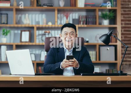 Asian boss works at a table in a classic office, types on the phone, looks at the camera and smiles Stock Photo