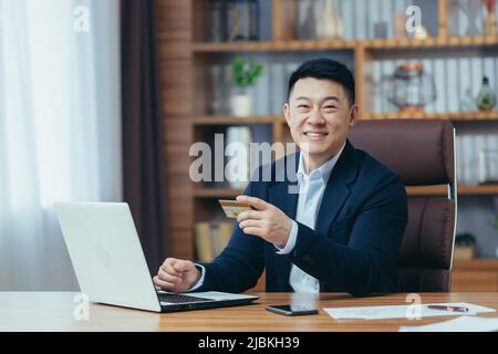 Young successful Asian man banker, banker holding a credit card, talking on a laptop on a video call, consulting, selling. looking at the camera, smil Stock Photo