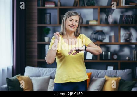Portrait of a senior smiling woman at home, doing fitness and looking at the camera watching the pulse on the fitness smart watch, happy with the resu Stock Photo