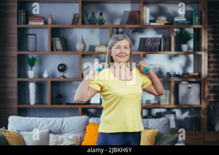 Portrait of senior smiling woman at home, doing fitness and looking at camera holding dumbbells, morning exercise Stock Photo