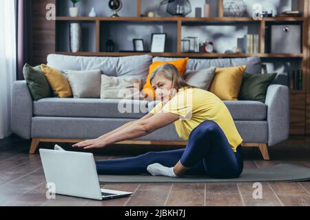 Senior woman doing fitness, sitting on the floor at home, using laptop for online sports group activities Stock Photo