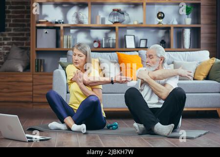 Senior couple, man and woman doing morning exercises together, doing fitness sitting on the floor, using laptop for online classes Stock Photo