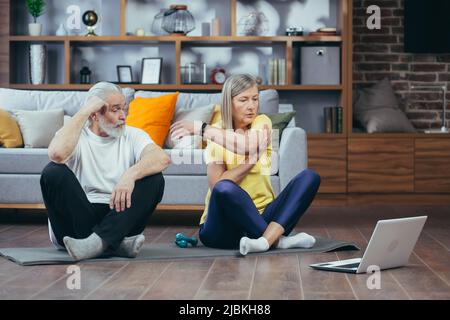 Senior couple, man and woman doing morning exercises together, doing fitness sitting on the floor, using laptop for online classes Stock Photo