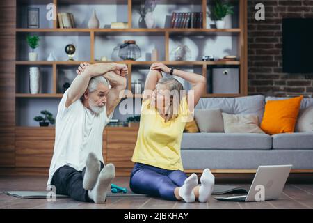 Senior couple, man and woman doing morning exercises together, doing fitness sitting on the floor, using laptop for online classes Stock Photo