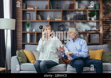 senior married couple, quarreling, gray-haired man and woman sitting on sofa, wife annoyed talking on the phone Stock Photo