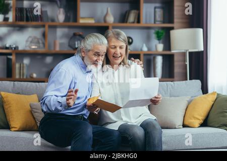 Happy senior retired couple sitting on sofa at home reading letter, happy and smiling Stock Photo