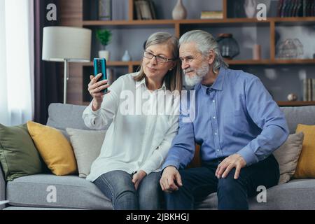 Elderly couple of senior retirees Happy gray haired grandparents communicate online in a video call using a mobile phone smartphone. laugh and wave ha Stock Photo
