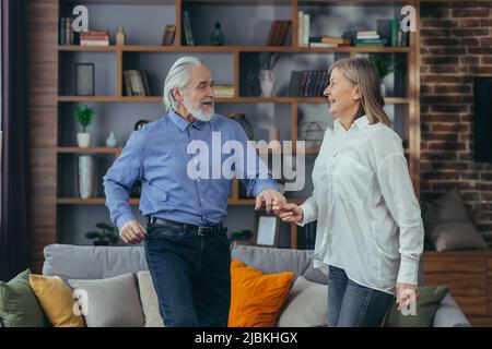 Happy cheerful senior married couple having fun at home. older family is dancing. Gray haired retirees rejoice moving active lifestyle. Old man and wo Stock Photo