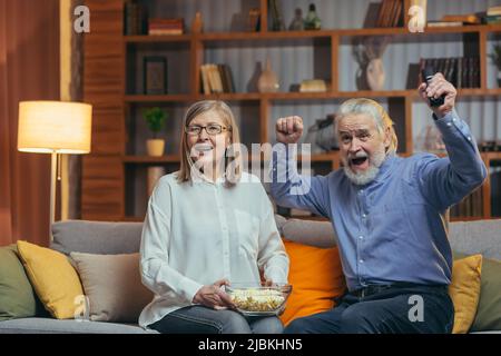 Happy senior couple cheers in watching sport competitions television TV game celebrating victory together on sofa in living room at home. mature aged Stock Photo