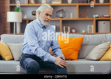 The older man is sitting on the couch at home, has pain in the knee joint, holding his hands and feet Stock Photo