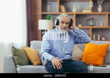 Senior gray-haired man sitting on sofa at home, relaxing and listening to music on headphones, using music app on phone Stock Photo