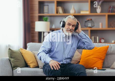 Senior gray-haired man sitting on sofa at home, relaxing and listening to music on headphones, using music app on phone Stock Photo