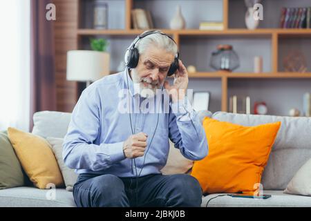 Senior gray-haired man sitting on sofa at home, relaxing and listening to music on headphones, using music app on phone Stock Photo