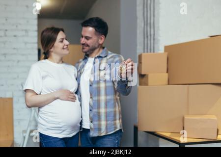 Young happy family expecting a baby. Pregnant woman and husband moved to a new house, apartment. Unpack boxes with things. Hugging, smiling Stock Photo