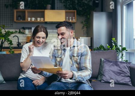 Young family. The man and the pregnant woman received a letter, documents, a mortgage permit, and a loan. Very happy. Sitting on the couch at home. Stock Photo