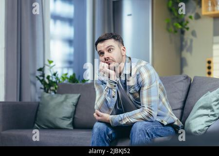 Portrait of a handsome young man. Waiting, bored, thought. Sits at home on the couch, holds his head in his hand, thinks depression, quarantine Stock Photo