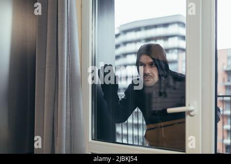 Young male robber in dark clothes looks out the window of the apartment, house. He wants to go into housing Stock Photo