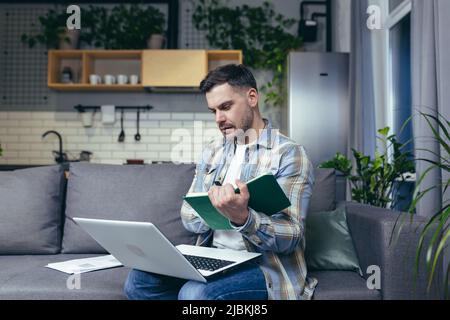 Distance learning online. Young man, student. He studies at home with a laptop, books. Sitting on the couch writes in a notebook online lecture. Liste Stock Photo