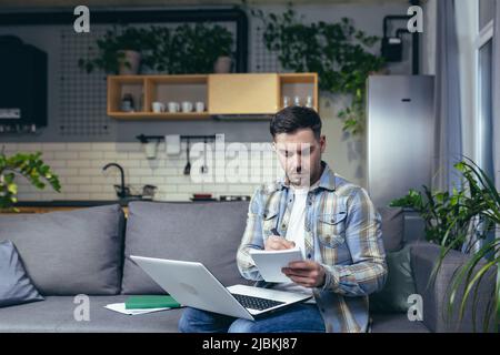 Distance learning online. Young man, student. He studies at home with a laptop, books. Sitting on the couch writes in a notebook online lecture. Liste Stock Photo