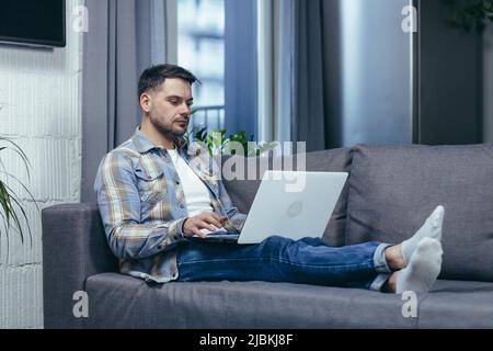 Freelancer. Handsome young man working on laptop at home. Lying on the couch in home clothes. Stock Photo