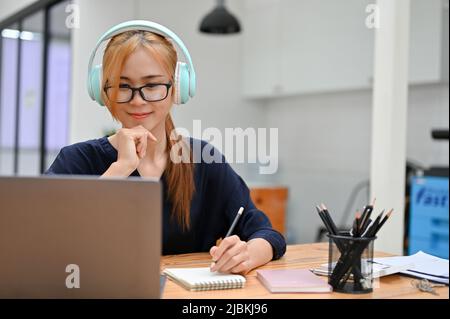 Happy Asian female student wearing headphones watch a webinar, listen to an online course study, and look at her laptop screen at home. Stock Photo