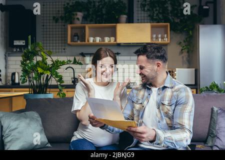 Pregnant woman at home with her husband received a letter with good news, young family sitting on the couch Stock Photo