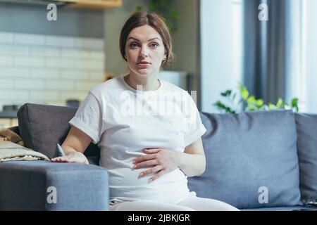 Pregnant woman at home sitting on the couch and talking on video call, listening to online doctor's consultation focused Stock Photo