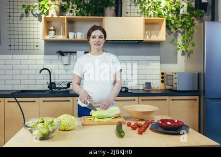 Pregnant woman preparing healthy food in the kitchen. He looks at the camera, smiles. chops vegetables and fruits. Stock Photo
