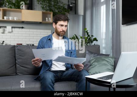 Pensive man at home working on laptop, freelancer on paper focused and serious Stock Photo