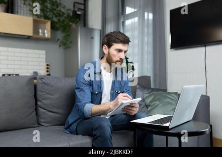 The man at home is studying remotely, uses a laptop, sitting at home on the couch, recording a lecture Stock Photo
