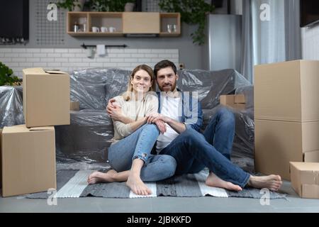 young family in a new apartment, unpacking things from cardboard boxes, man and woman sitting together in love on the floor smiling and looking at the Stock Photo