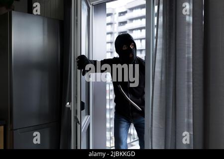 A male thief in a black mask, enters the apartment for robbery, the robber broke the door lock Stock Photo