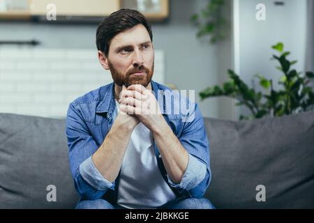 The man at home is sitting on the couch, resting looking away, smiling dreamy pensive Stock Photo