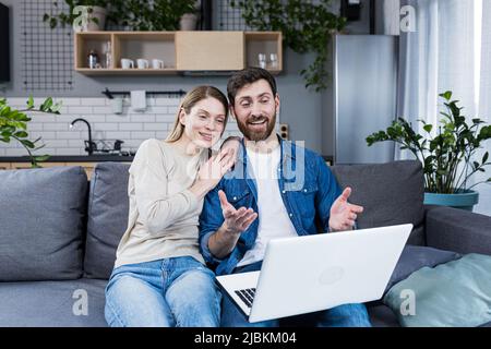 Married couple man and woman sitting together at home on the couch using laptop for video chat and chatting with friends Stock Photo