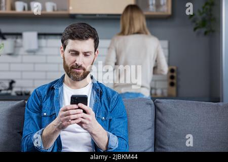 Close up photo. Conflict, young couple, family, man and woman. The man sits on the couch, looks at the phone, plays games, does not pay attention to t Stock Photo