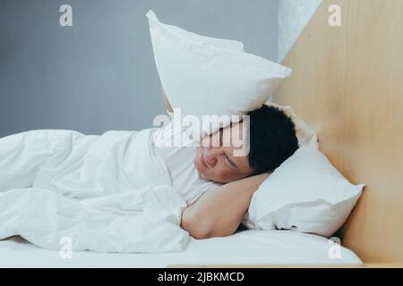 A man at home tries to fall asleep, noisy neighbors interfere with sleep, an Asian closes his ears with pillows, tired after work Stock Photo