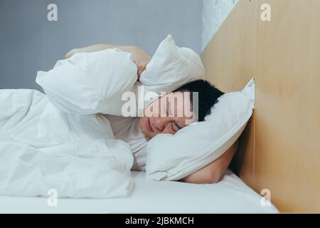 A man at home tries to fall asleep, noisy neighbors interfere with sleep, an Asian closes his ears with pillows, tired after work Stock Photo