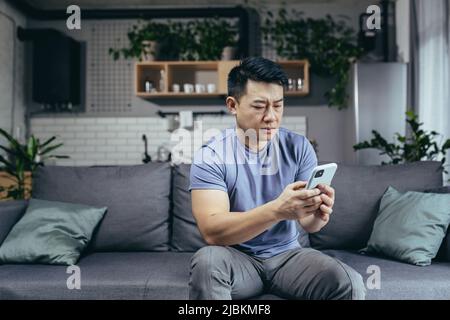 Man reading bad news from phone, asian frustrated and sad looking at phone screen sitting on sofa at home Stock Photo