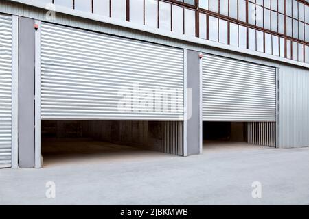 Opening gray iron shutter door of garage and industrial building warehouse exterior facade with grey concrete road, side view nobody. Stock Photo