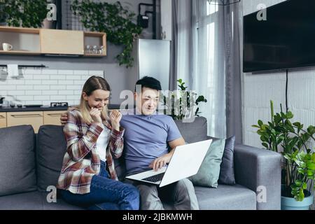 Happy young asian couple bonding laughing using laptop sit at table home Stock Photo